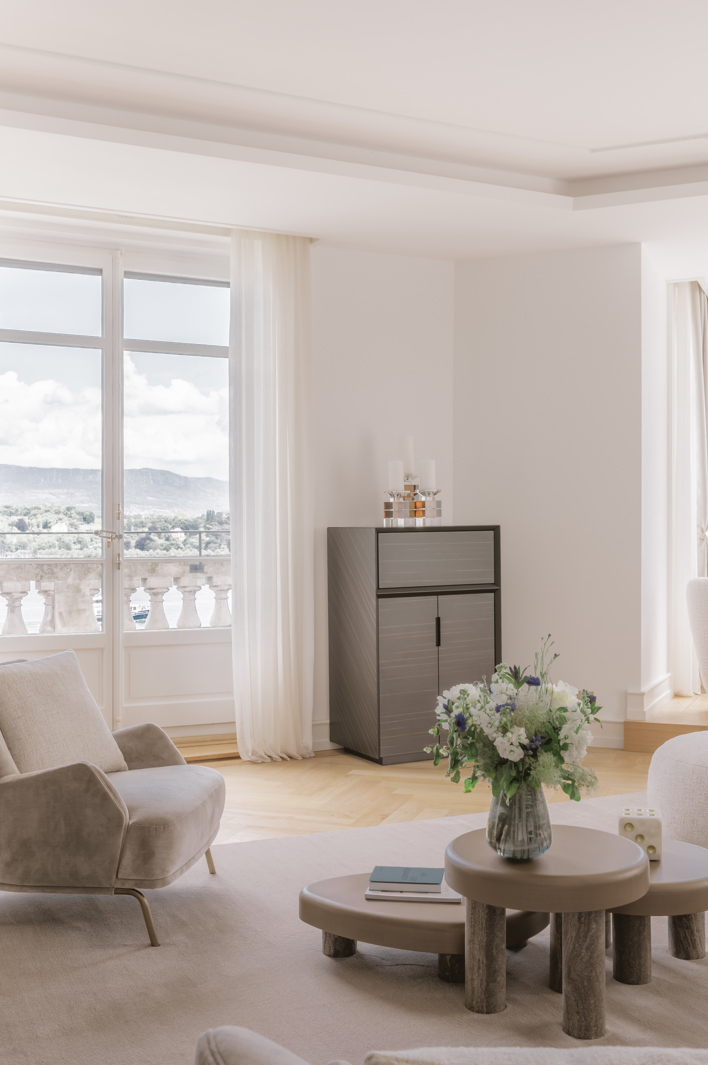 Bright modern living room with minimalist décor, light furniture, large windows overlooking a balcony, and soft natural lighting.