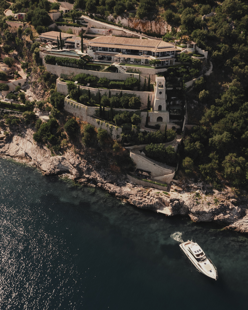 منظر جوي لفيلا فاخرة على منحدر صخري محاطة بالخضرة الكثيفة ويبحر بجانبها يخت على الساحل الصخري.