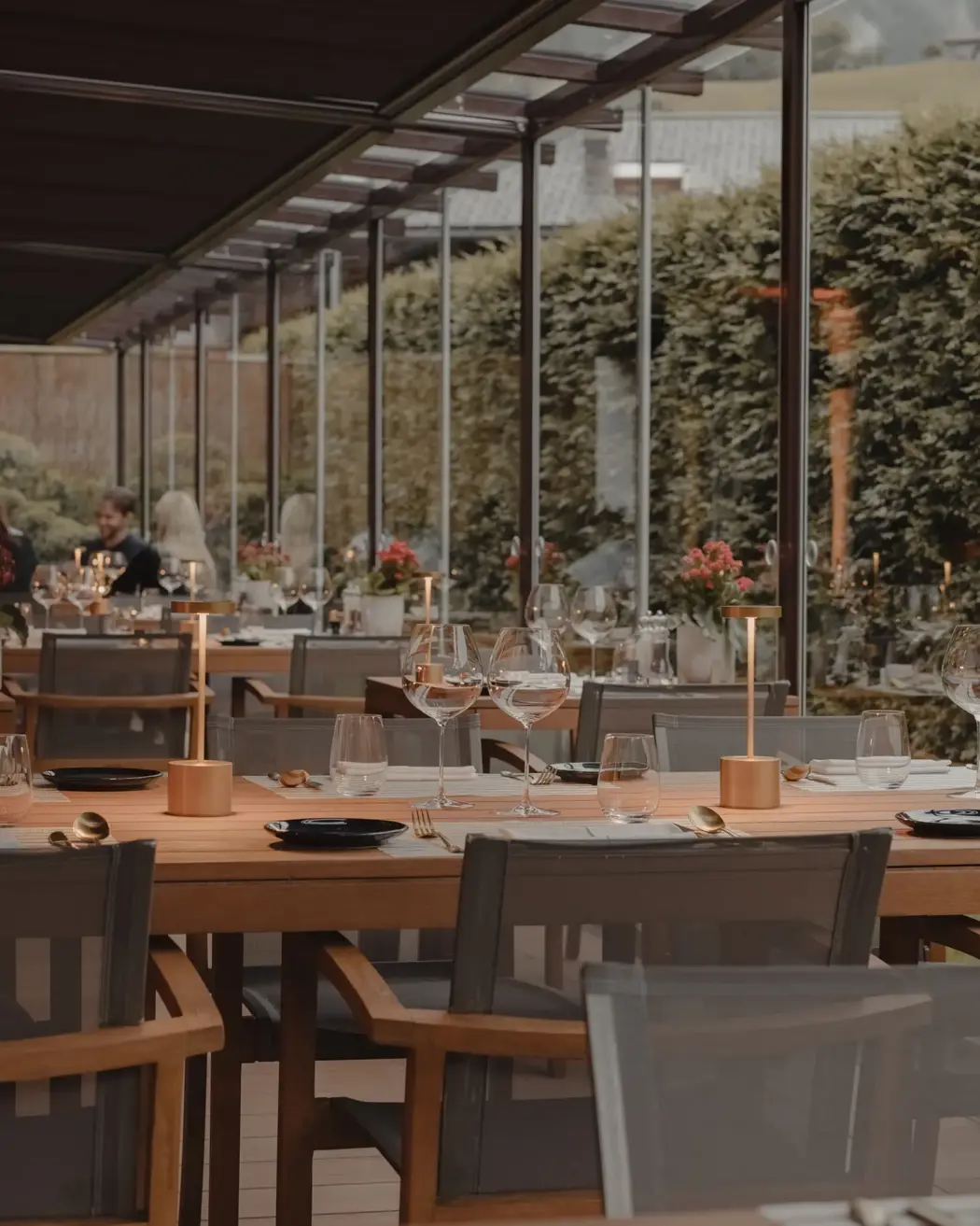 An elegant restaurant dining area with glass walls and wooden tables set with wine glasses, candles, and flowers, creating a warm and refined ambiance with guests seated in the background.